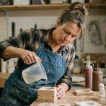 Femme versant du silicone liquide dans un moule en bois sur une table d'atelier bricolage maison, avec des outils de moulage décoratif en arrière-plan
